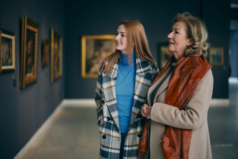 Grandmother and adolescent granddaughter are looking at the paintings in the art gallery.
