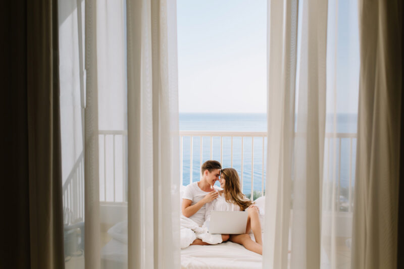 Portrait of loving couple watching movie together on balcony in morning with curtains on foreground. Young man resting with his amazing tanned girlfriend in bed holding laptop with sea background.
